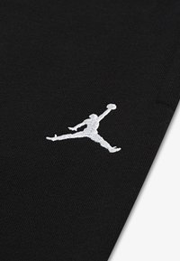 Juodas audinys su baltu išsiuvinėtu Jumpman logotipu. Paviršius lygus, artimas žvilgsnis akcentuoja siuvimo detales.