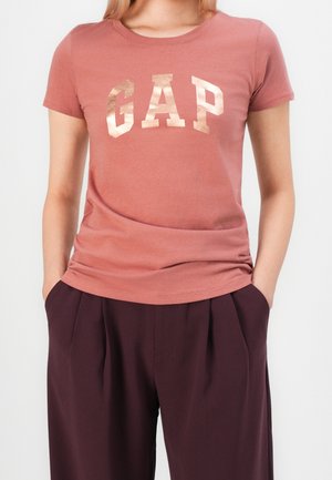 Kvinde iført en støvet lyserød GAP-logo t-shirt med skinnende bogstaver og løstsiddende mørkelilla plisserede bukser, hænderne i lommerne.