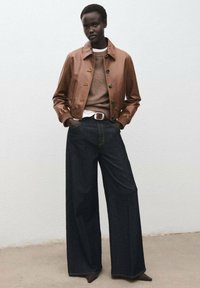 Giacca di pelle marrone con bottoni davanti, maglione beige a maglia, classica t-shirt bianca e jeans in denim scuri a gamba larga. Una cintura semplice accentua la vita.
