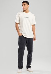 T-shirt en coton crème avec le texte "NEW YORK" en vert, associée à un jean noir. Le modèle porte des baskets blanches et se tient devant un fond neutre.