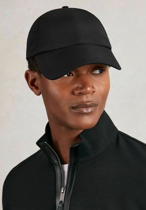 Sort baseballcap med buet skygge, sømløst design og syninger; iført med en sort zip-jakke med høj krave.