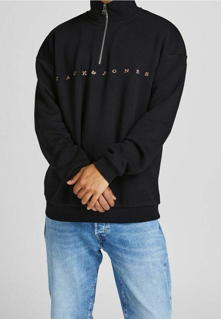 Personne portant un sweat-shirt noir Jack & Jones à fermeture éclair quart de zip, les mains jointes, associé à un jean bleu clair sur un fond uni.