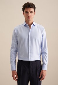 Camicia a quadri blu chiaro con colletto a punta, maniche lunghe e bottoni bianchi, abbinata a pantaloni scuri. Tessuto liscio, vestibilità sartoriale.
