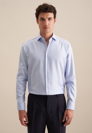 Camicia a quadri blu chiaro con colletto a punta, maniche lunghe e bottoni bianchi, abbinata a pantaloni scuri. Tessuto liscio, vestibilità sartoriale.