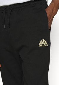 Black sweatpants made of soft fabric, featuring an elastic waistband with drawstring. Gold embroidered Fila logo in a triangular shape on the thigh.