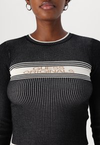 Maglione nero a costine con silhouette sagomata, caratterizzato da una banda color crema e dal logo "GUESS ORIGINALS" in lettere dorate sul petto.