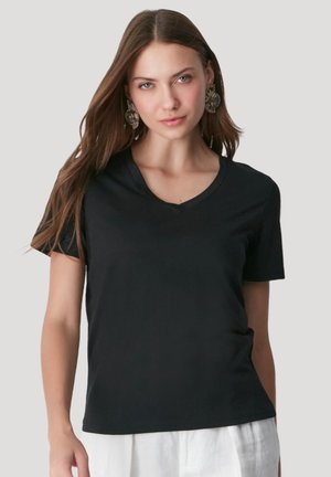 Woman with long brown hair wearing ornate earrings, a black V-neck T-shirt, and white pants against a plain light gray background.