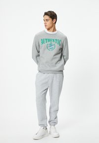 Grå sweatshirt med "AUTHENTIC" tekst i grøn, parret med matchende grå sweatpants og hvide sneakers. Casual pasform og blød tekstur.