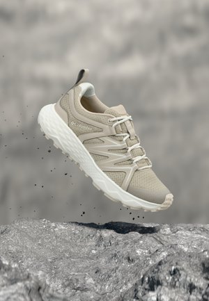 Beżowy but do wędrówek Columbia z białą podeszwą i sznurowadłami, przedstawiony w powietrzu nad kamienistą powierzchnią z unoszącymi się cząstkami kurzu.