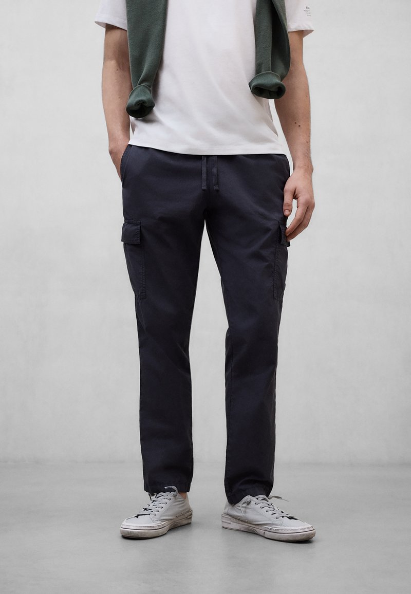 Hombre con pantalones cargo oscuros, zapatillas blancas, camiseta blanca y un suéter verde sobre los hombros, de pie frente a un fondo gris claro.