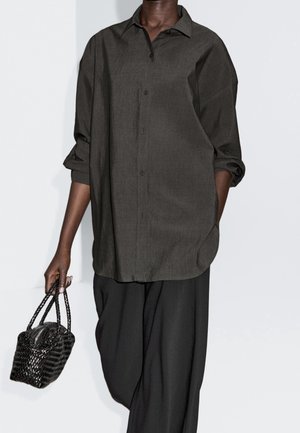 Osoba ubrana w luźną, ciemnoszarą koszulę zapinaną na guziki z podwiniętymi rękawami oraz czarne, szerokie spodnie, trzymająca czarną plecioną torebkę.
