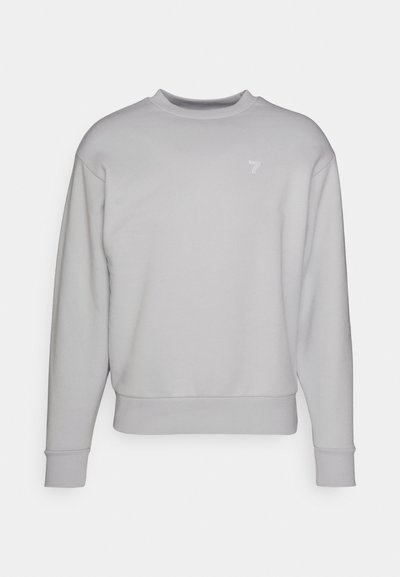 Sudadera gris claro con cuello redondo, mangas largas y puños acanalados. Presenta un sutil logo blanco en el lado izquierdo del pecho. Textura de tela suave.