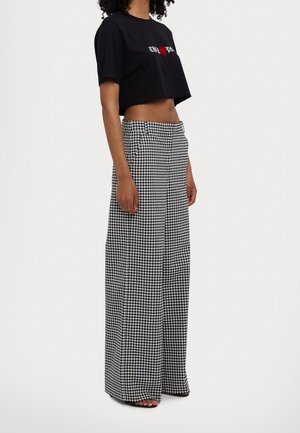 Femme portant un t-shirt noir court avec un imprimé cœur rouge et un pantalon large taille haute à carreaux noir et blanc sur fond blanc.