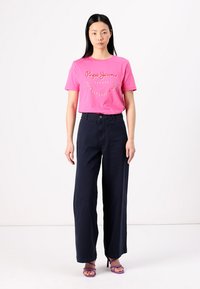 T-shirt de algodão rosa com logótipo bordado, combinado com calças largas azul-marinho e sandálias roxas de salto alto, apresentando uma textura suave e um corte casual.