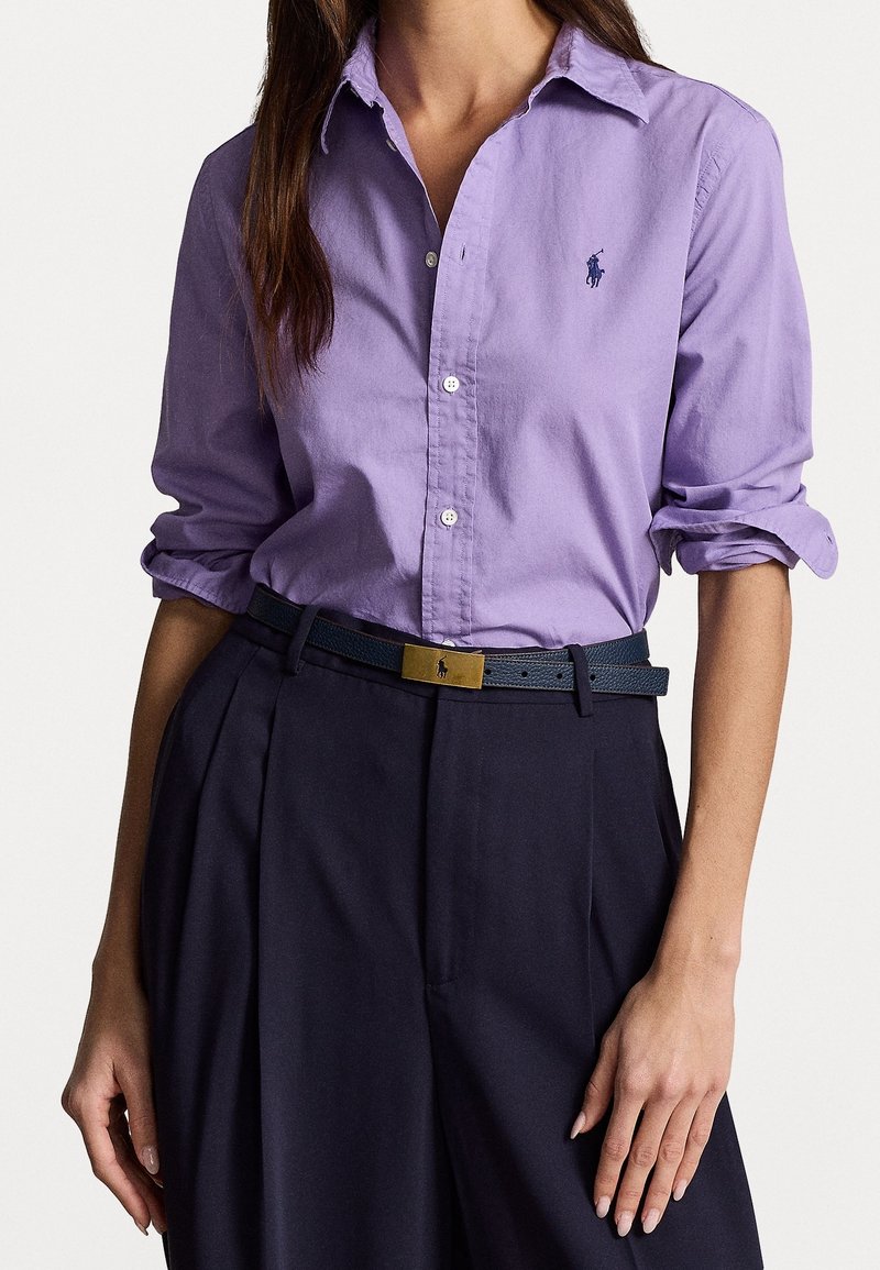 Camisa de manga larga color púrpura claro con botones, con un pequeño logo bordado. Combinada con pantalones azul marino oscuro y un cinturón negro texturizado con hebilla dorada.