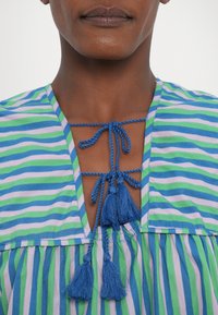 Top a righe blu e verdi con scollo a V, dotato di chiusura con nodo e due frange blu. Il tessuto appare liscio con una consistenza leggera.
