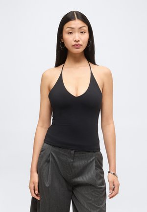 Jeune femme aux longs cheveux noirs portant des boucles d'oreilles créoles, un haut dos nu noir et un pantalon large gris à fines rayures, debout devant un fond clair.