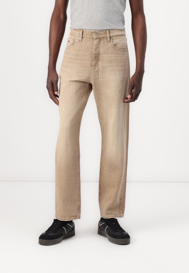 Beige denim jeans with a straight cut, five pockets, and a visible logo on a front pocket. Worn with black sneakers.