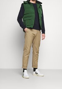 Vihreä toppaliivi hupulla, tummansininen pusero, ruskeat kapenevat housut ja valkoiset lenkkarit mustilla yksityiskohdilla. Rentoon tyyliin sopiva, kerroksittain yhdistetty asukokonaisuus.