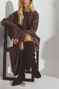 Un pull marron à manches côtelées associé à une jupe marron, accompagné de bottes noires en daim jusqu'au genou avec un détail de fermeture éclair sur le côté.