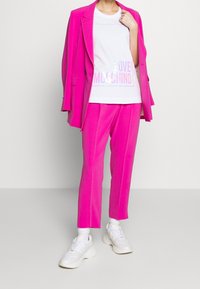 Světle růžový oblek s přizpůsobeným sakem a kalhotami, doplněný bílým tričkem s nápisem "LOVE MOSCHINO" v pastelových barvách, dokončeným bílými teniskami.