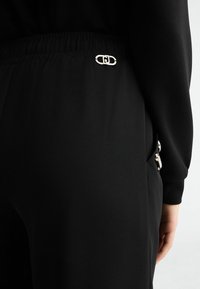 Zbliżenie na czarne spodnie z elastycznym pasem i srebrną ozdobną sprzączką, noszone z czarną bluzką z długim rękawem, a ręka spoczywa obok biodra.