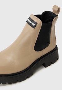 Botín Chelsea de cuero beige con paneles elásticos laterales negros, un tirador y una suela de goma negra resistente con surcos profundos.
