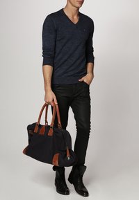 Homme portant un pull en V bleu foncé, pantalon noir, bottes noires, tenant un sac noir avec des poignées et des accents en cuir marron dans la main droite.