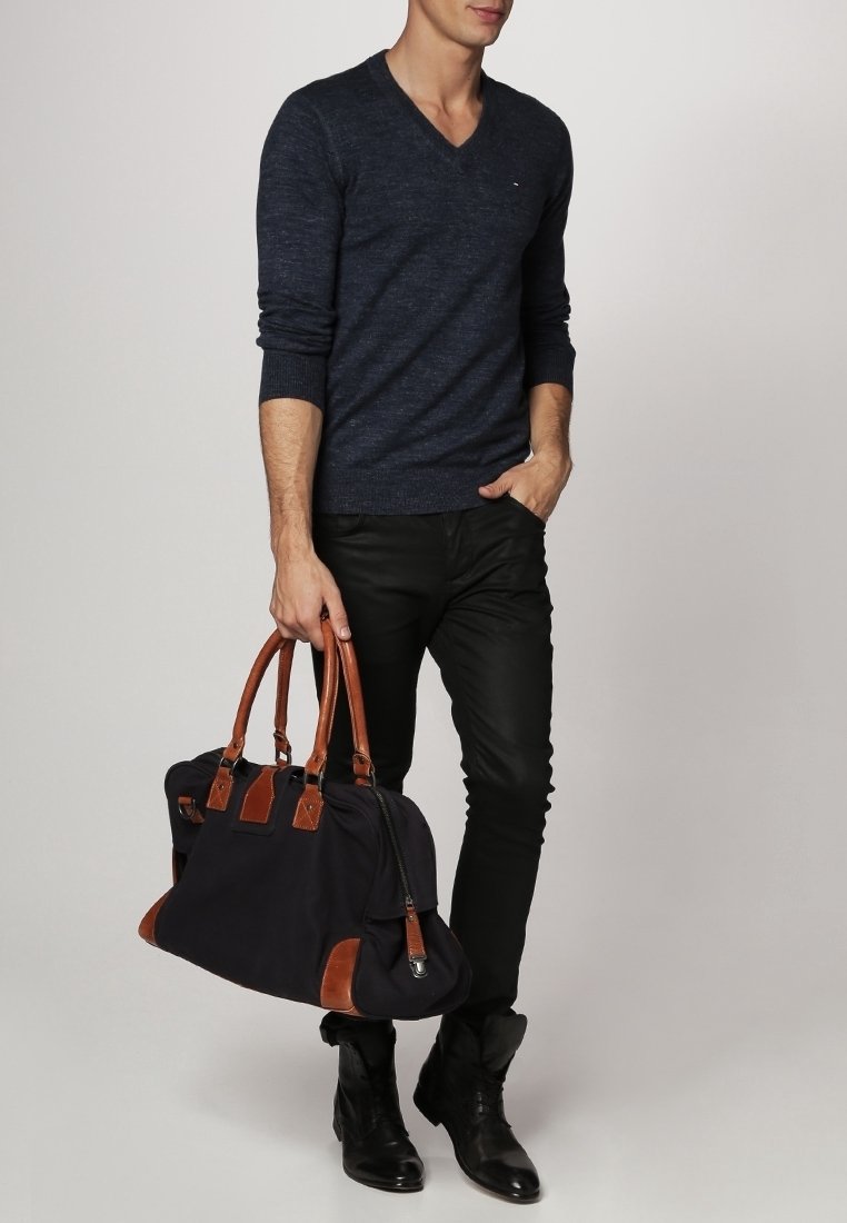 Homme portant un pull en V bleu foncé, pantalon noir, bottes noires, tenant un sac noir avec des poignées et des accents en cuir marron dans la main droite.