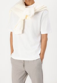 Man in een wit T-shirt met korte mouwen en lichtgrijze sweatpants met een crème sweatshirt om zijn schouders gedrapeerd. Handen in de zakken.