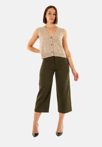 Gilet en tricot sans manches beige clair avec fermeture à boutons, associé à un pantalon large taille haute vert olive et des talons noirs.