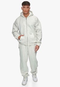 Haut à capuche zippé en polaire gris clair avec poches передні, associé à un pantalon de jogging assorti ; les deux présentent une texture douce et une coupe décontractée.