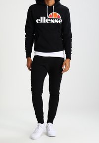 Svart hoodie med vitt logotyp som har en regnbågsbåge, i kombination med svarta åtsittande joggers och vita sneakers. Kontinuerliga dragstrings vid huvan.