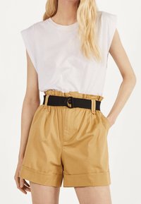 White sleeveless top with shoulder pads and high-waisted beige shorts, featuring a black belt with a round buckle and side pockets.