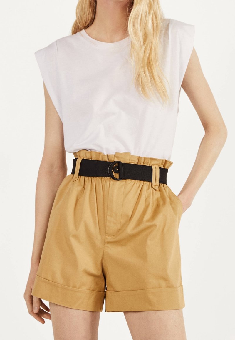 White sleeveless top with shoulder pads and high-waisted beige shorts, featuring a black belt with a round buckle and side pockets.