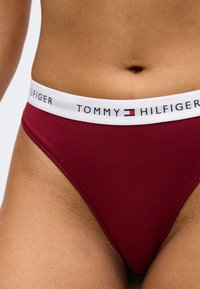 Burgundijas kokvilnas stringi ar baltu elastīgu jostu, uz kuras redzams "TOMMY HILFIGER" logo, izceļoties ar gludu tekstūru un pieguļošu dizainu.