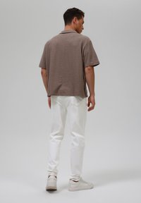 Polo shirt corto marrone a maniche corte con una finitura texturizzata, abbinato a jeans bianchi e sneakers chiare, visto da dietro.