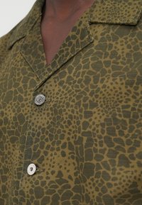 Πουκάμισο με κοντά μανίκια σε ελιά πράσινο ύφασμα με υφή ζωικής εκτύπωσης, διαθέτει γιακά με εγκοπή και δύο κουμπιά από ασημένιο μέταλλο.