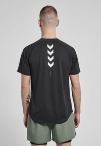 Sort atletisk t-shirt med korte ærmer, der har et hvidt chevronmønster på ryggen. Stoffet har en glat tekstur og atletisk pasform.