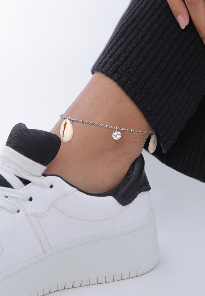 Sølv kæde ankelkæde med en hvid skal og et tekstureret rundt charm, båret ovenfor en sort og hvid sneaker, mod en neutral baggrund.