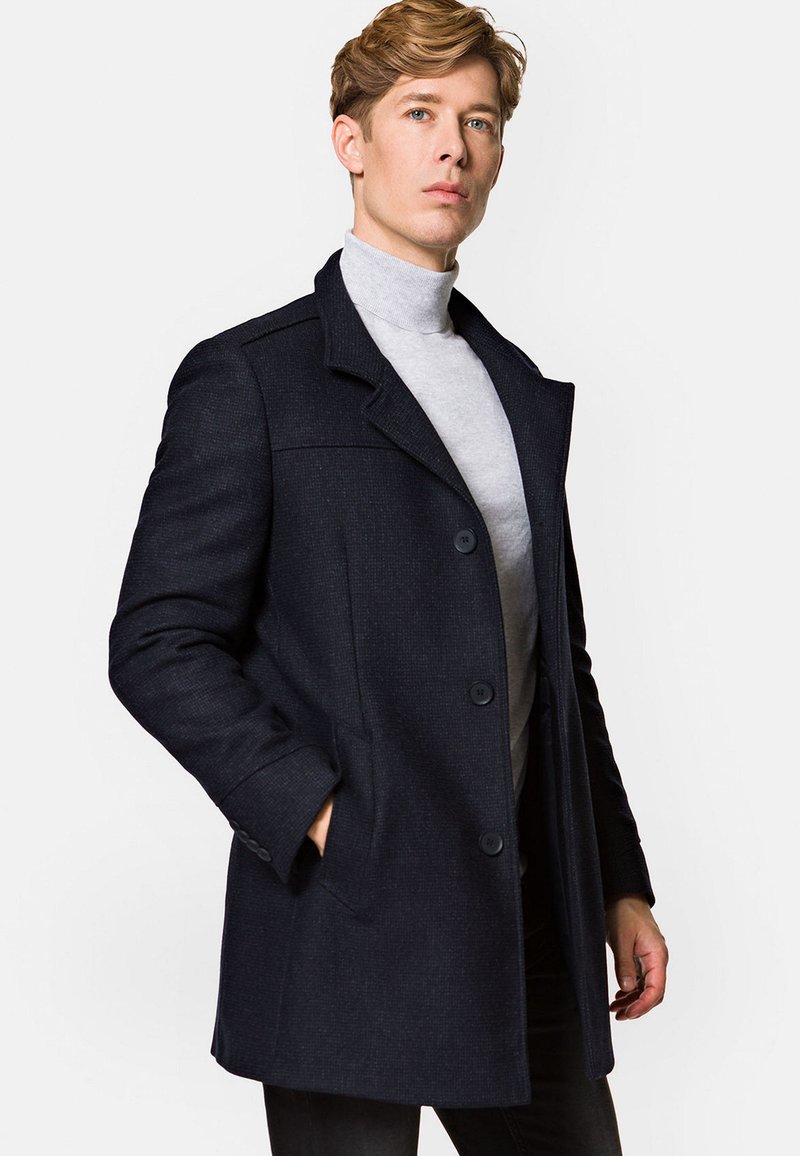 Granatowy płaszcz z wełny z domieszką, z wyraźnym kołnierzem, dwoma guzikami i bocznymi kieszeniami. Noszony na jasnoszary golf, podkreślający dopasowany fason.