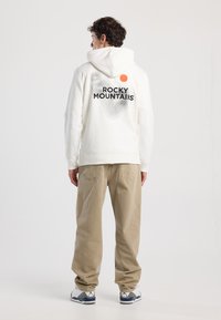 Felpa bianca con una grande grafica nera "ROCKY MOUNTAINS" e un punto arancione sul retro, abbinata a pantaloni beige e sneakers grigie.
