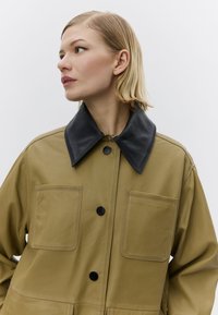 Khaki leren jas met een grote zwarte kraag, voorzien van twee voorzakken en zwarte knopen, met een gladde textuur en een gestructureerd ontwerp.