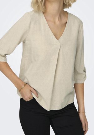 Kvinde iført beige v-hals, 3/4 ærmet bluse med frontplissering, sorte bukser samt gyldne armbånd og ringe, hånd i lommen på hvid baggrund.