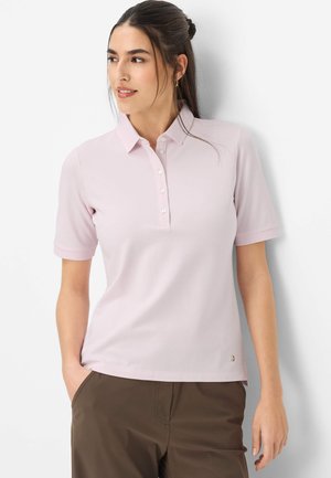 Kvinde med mørkt hår sat op i hestehale, iført en lyserød kortærmet skjorte med krave og brune bukser, stående foran en hvid baggrund.