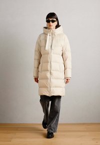 Crème-kleurige pufferjas met capuchon, front-rits en opvallende horizontale stiksels. Draag met donkere grijze wijde broek en zwarte schoenen.
