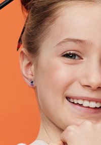 Fioletowe kolczyki w kształcie jednorożca z srebrnymi akcentami, noszone przez dziewczynę na pomarańczowym tle, ukazujące zbliżenie jej uśmiechniętej twarzy.