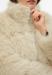 Beežist kunstkarvase mantliga, millel on kõrge krae, täiesti lukustatav tõmblukk ja pehme, tekstuuriga pind. Tuntud detailideks on õhuliselt tikitud paksud alad.