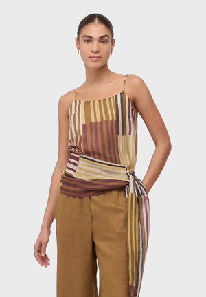 Femme portant un haut sans manches rayé avec un nœud sur un côté et un pantalon large couleur moutarde, debout avec une main dans la poche.