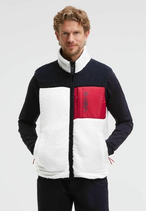 Man wearing a fleece jacket with black shoulders, white body, and a red chest pocket with "GA 97" text, hands in pockets, against white background.